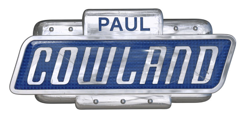 The Paul Cowland logo formed as a silver metal badge with blue ename inserts