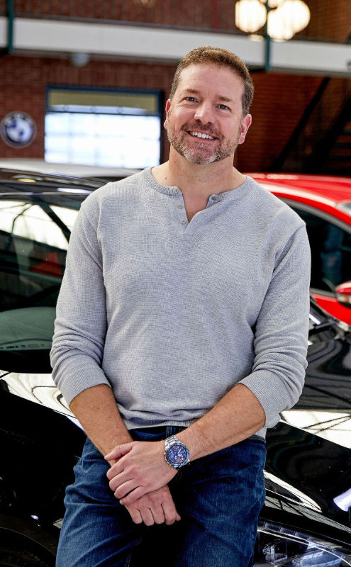 Paul leaning on the wheel arch of a blak Peugeot, with a red Ford and two other cars in the backgound. He has his hands crossed at his waist and a welcoming expression.